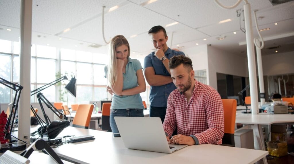 Two college students look over person typing on a laptop in an office with several desks and chairs