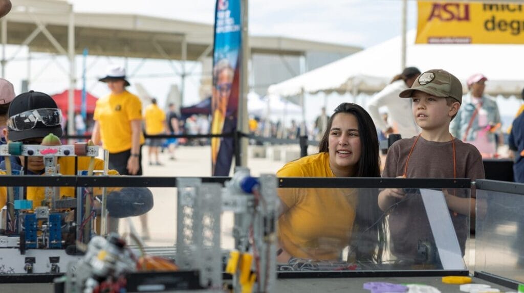 A person squatting down next to a child who is holding a controller of a robot at an ASU event