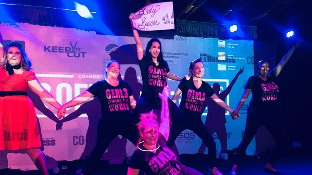 Six people on a stage with colorful lights holding a sign with text that reads ‘Lady Bosses #1’