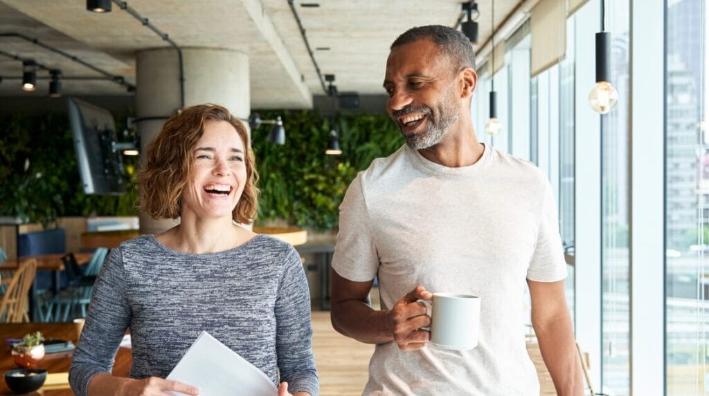 Two people smiling and walking in an office with a wall of windows on one side