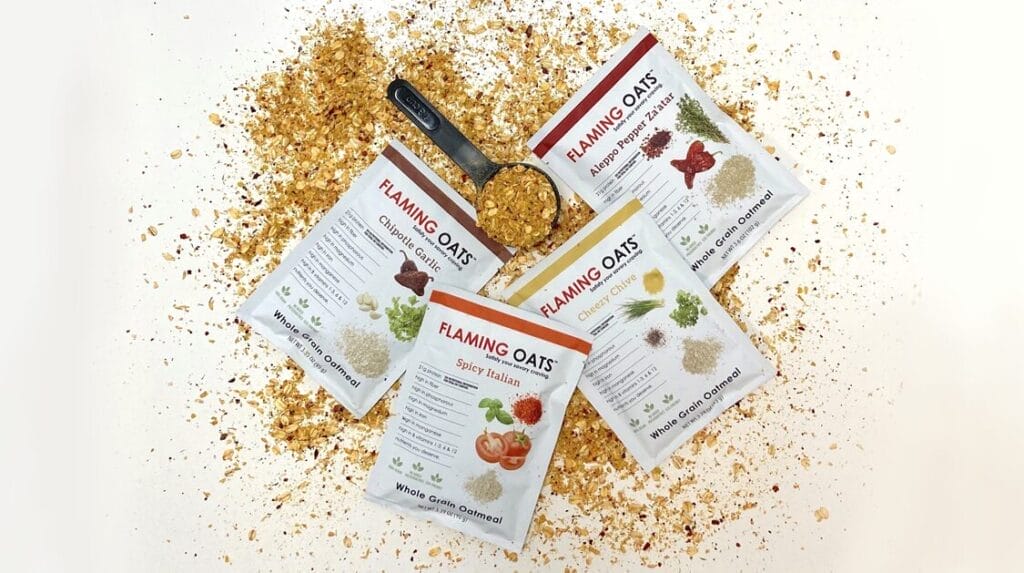 Four packets of Flaming Oats in a variety of flavors laying on top of a pile of oats and a scooper filled with oats next to the packets