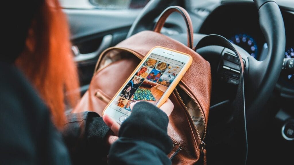 A person sitting in a car with a backpack leaning against the steering wheel with a mobile phone in their hand looking at a social media feed.