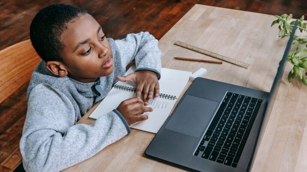 A child sitting at a table with his hands on a notebook and looking at a laptop screen.