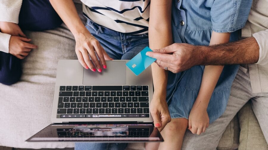 Two children and two parents sitting on a couch holding a credit card and typing on a laptop.