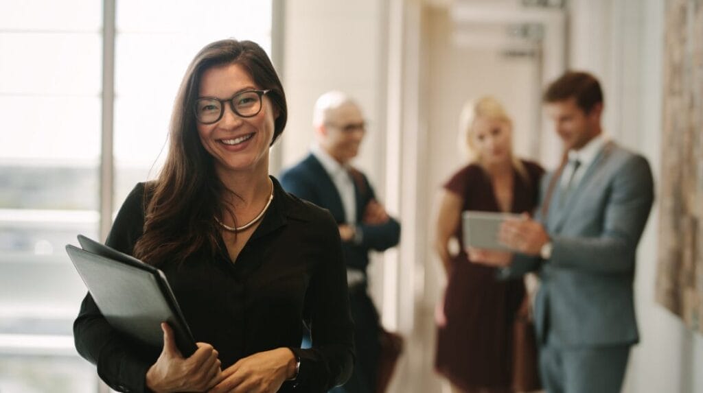 A person smiling and holding a binder with three business colleagues in the background having a conversation.