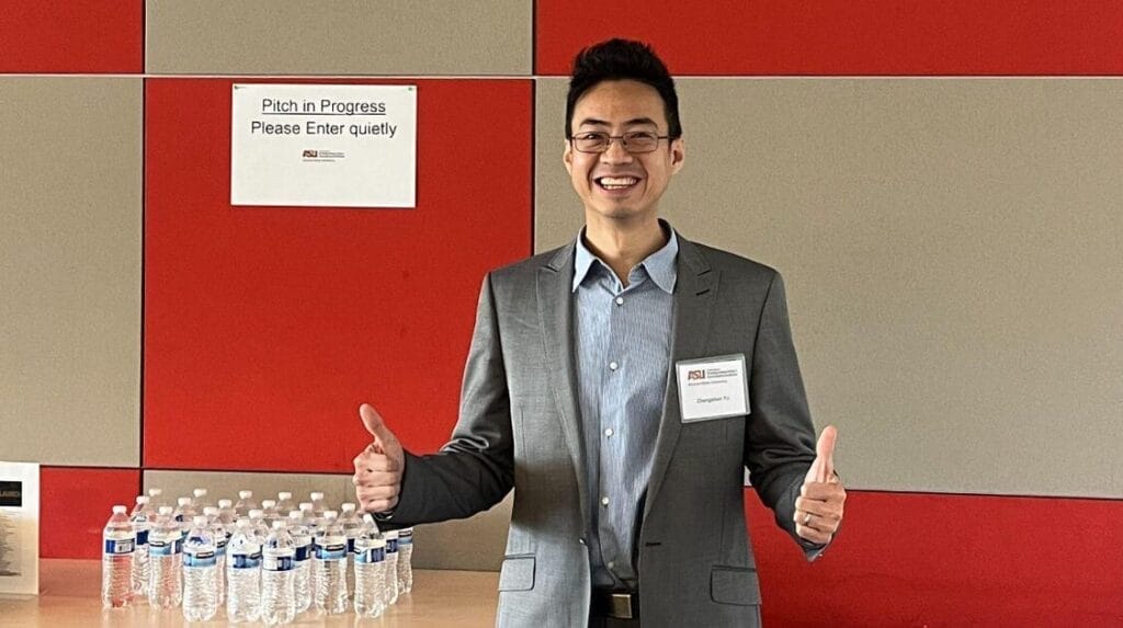 A person smiling with his two thumbs up with a table of water bottles next to him.