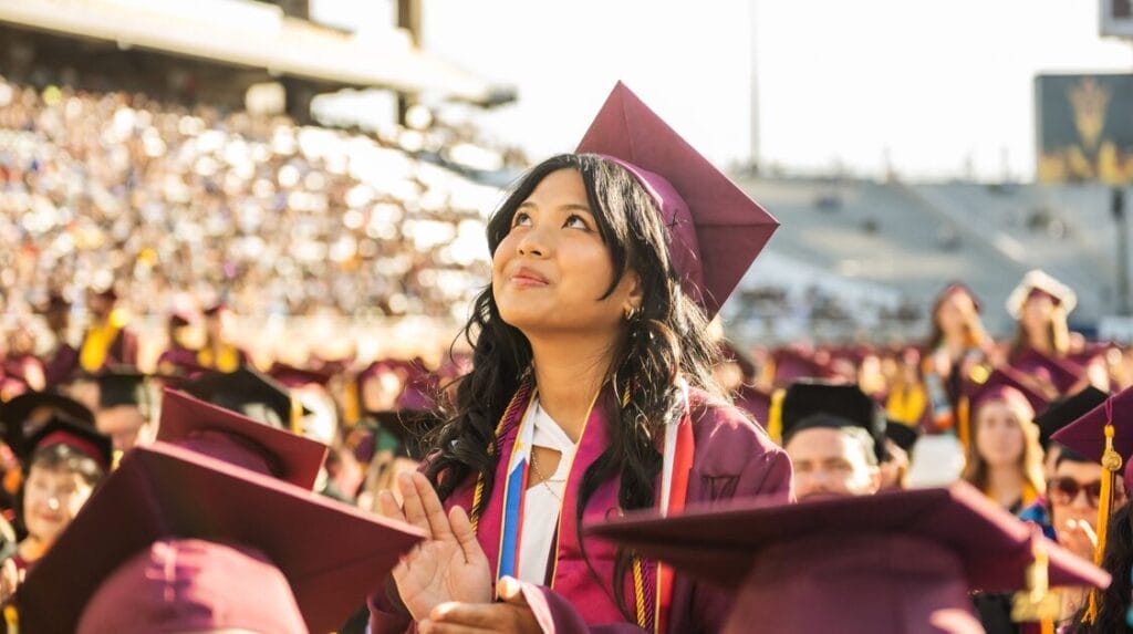 A person standing in a crowd, wearing a cap and gown, looking up at a college graduation.