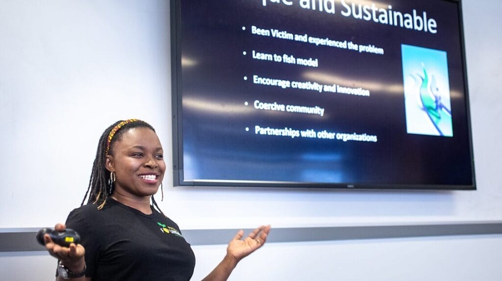 A person smiling and presenting her startup to a panel and audience with a screen showing her pitch deck