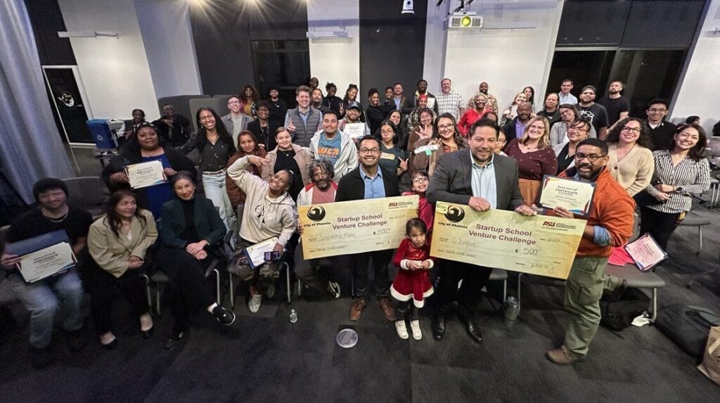A group of people in a coworking space smiling at the camera and two people holding large checks