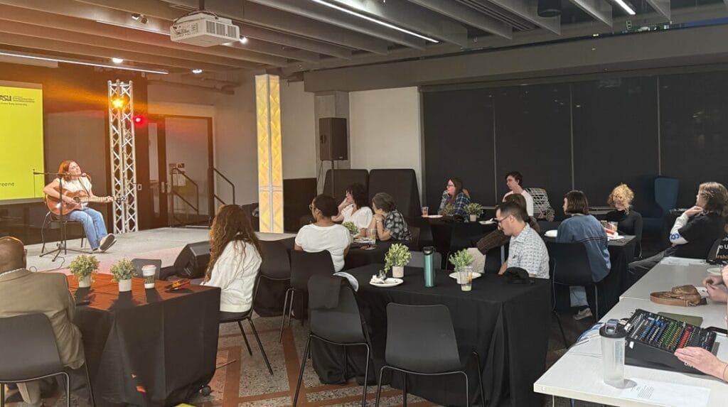 A college student sitting with a guitar on a stage and singing in front of an audience.