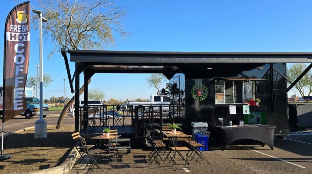 A coffee truck parked in a parking lot with two tables and chairs set in front of the truck.
