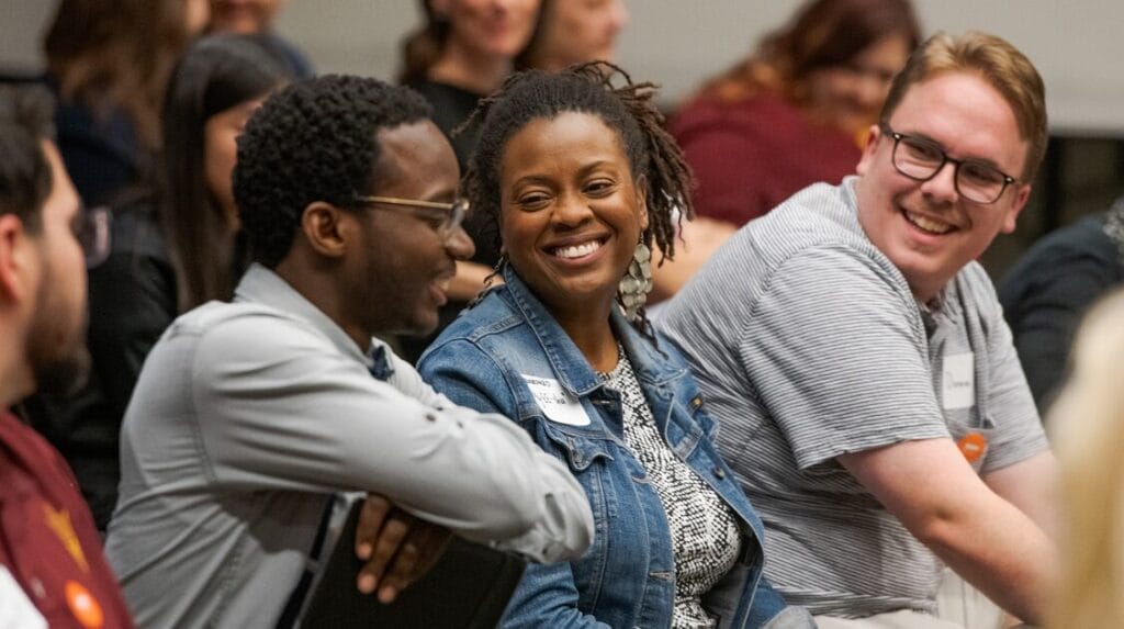 Three people smiling a talking while sitting in a crowd at an indoor event.

