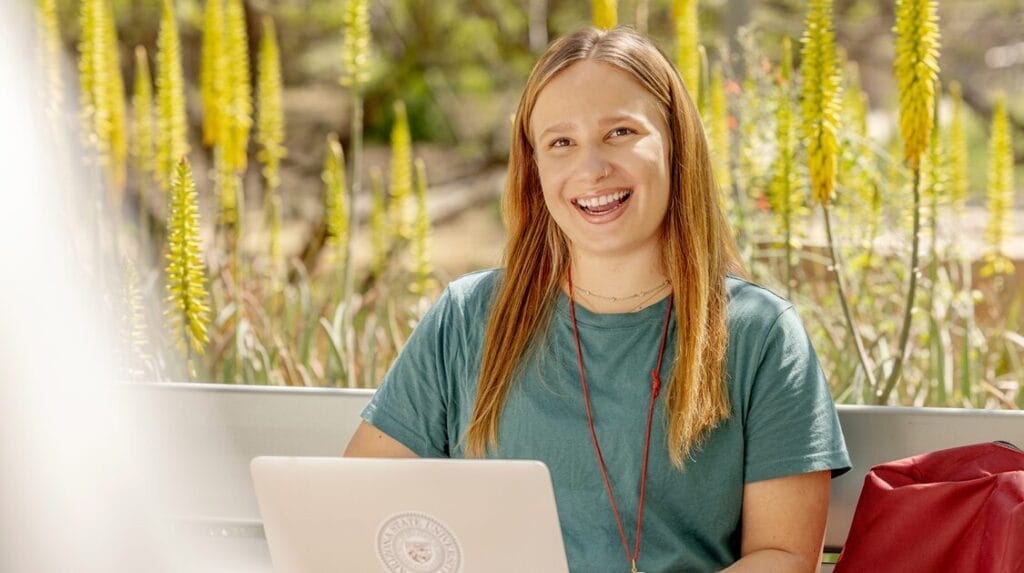 A person sitting outside smiling and working on her laptop.