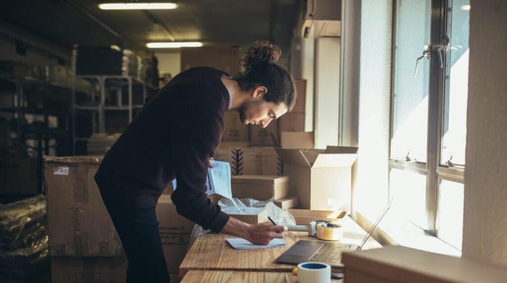  A business owner working in his warehouse, writing on a pad of paper next to a laptop.