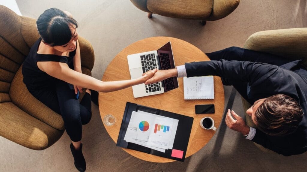 Top-down view of two people shaking hands at a table with a laptop, paperwork, coffee, and a phone.