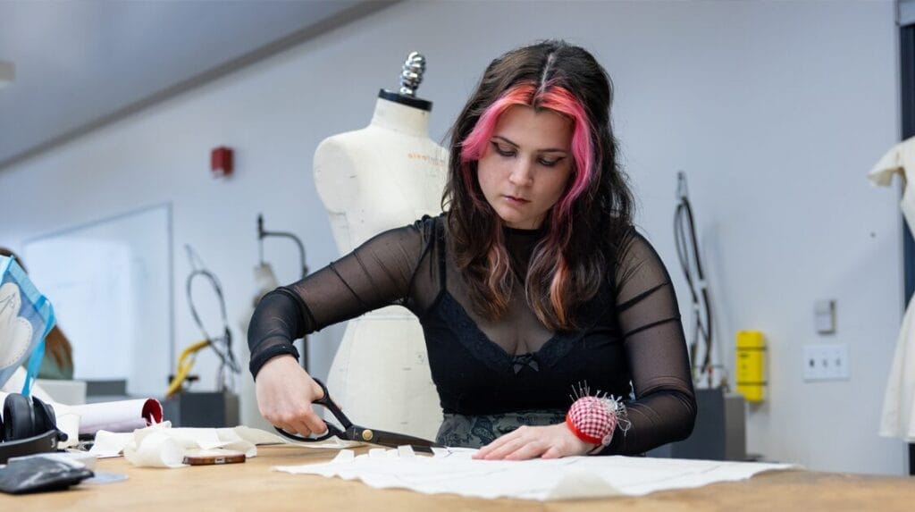 A college student standing at a table cutting a clothing pattern with dress forms in the background
