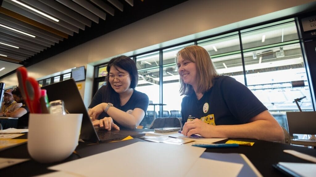 Two people sit at a table and look at the screen of a laptop