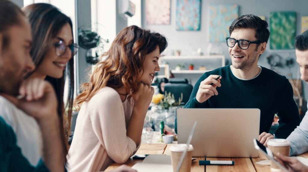 Five coworkers gather around a table, smiling as one teammate leads the conversation with his laptop open in front of him.
