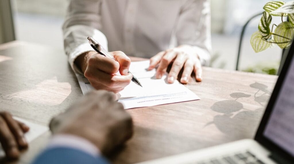 A person’s hand signing paperwork with another person’s hands in the foreground. 