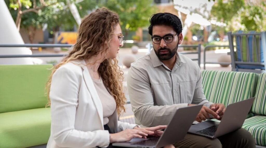 Two people sitting on an outdoor sofa looking at each other with laptops open on their laps