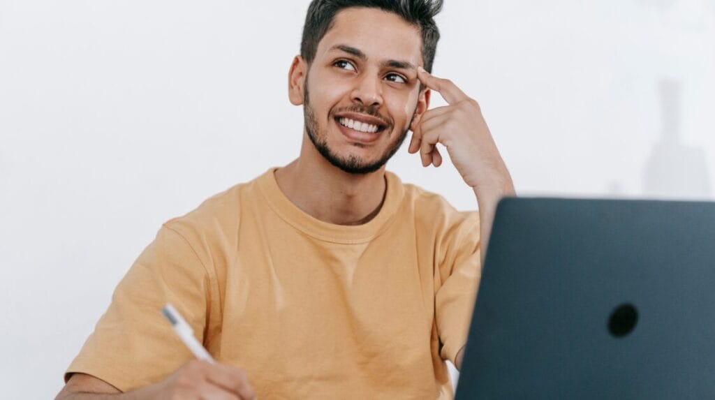 A person thinking, smiling and sitting at a desk with a laptop and a pen.
