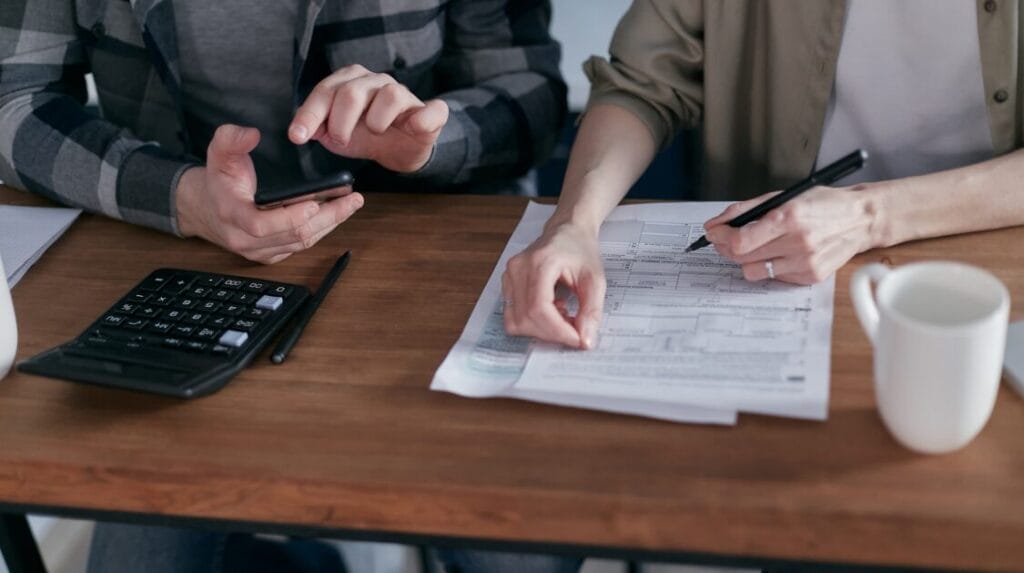 Two coworkers sitting at a desk looking at paperwork and one person tapping on a calculator.
