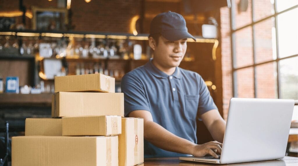 A person sitting in their office typing on a laptop with shipping boxes off to the side.