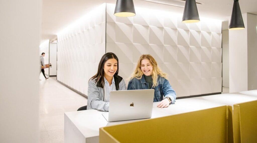 Two people sitting at a desk smiling and typing on a laptop.