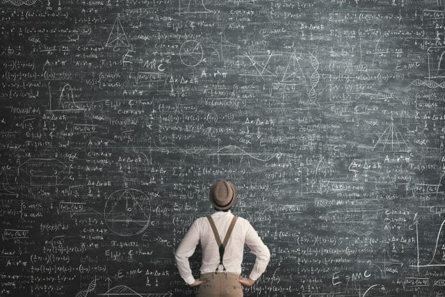 Man facing a chalkboard full of text. The chalkboard is much taller than the man. Only the back of the man can be seen.