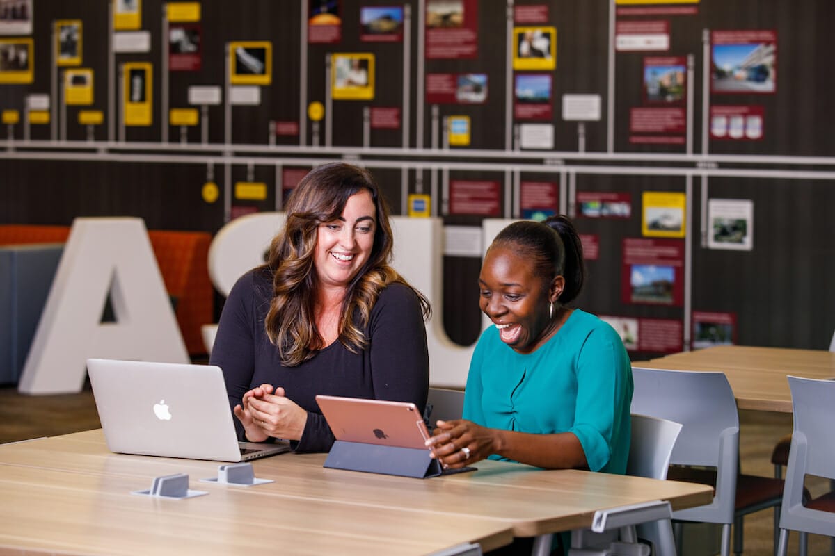 Two women sitting at a table with laptops. Both women are smiling