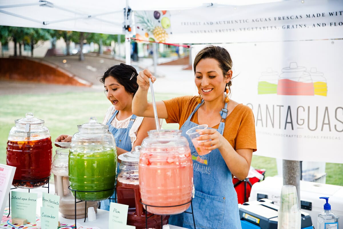 Two women, one is ladling a drink into a cup from one of two large pitchers