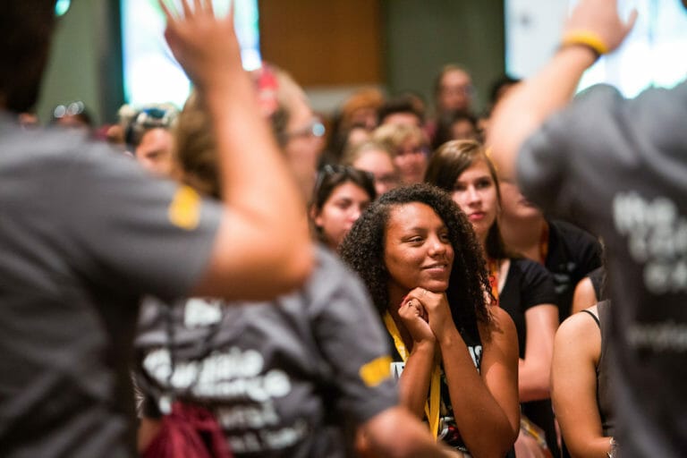 In a crowd of people, one women is in focus. Her hands are clasped below her chin.
