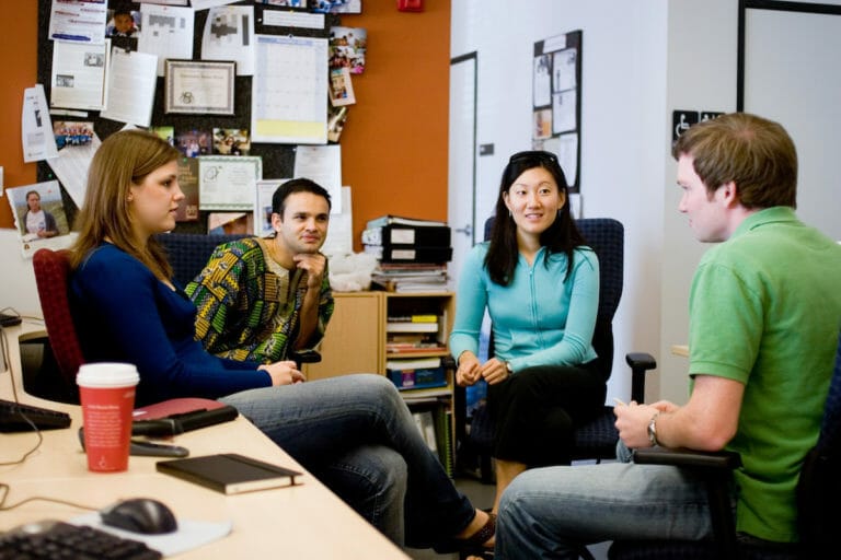 Four people sitting in chairs in an office, looking at each other, talking