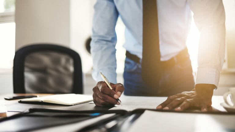 Young African entrepreneur wearing a shirt and tie leaning on his desk in an office signing documents