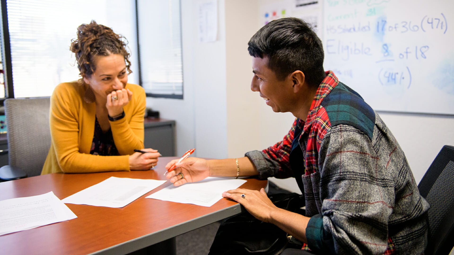 Woman and man sitting across from each other at a desk. The woman has her hand over her mouth thoughtfully, and is looking at the man. The man is holding a pen and looking down at his paper