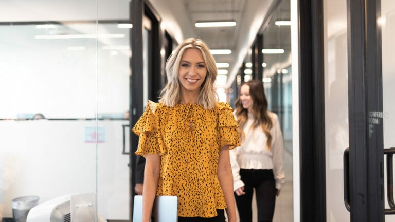 A women is standing in a hallway, holding a laptop, smiling