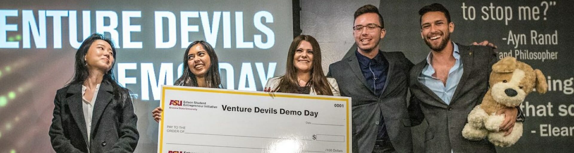Five people smiling and standing on a stage in dress clothes holding a large Arizona State University Venture Devils Demo Day check with a ‘Venture Devils’ sign in the background.